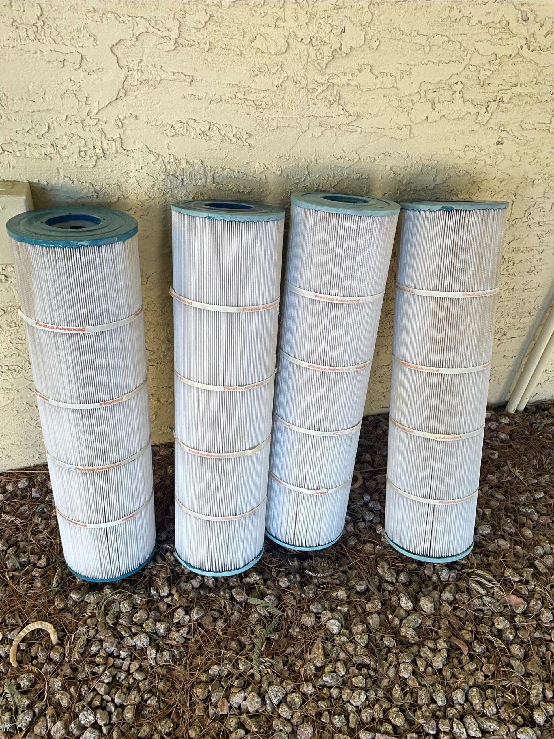 Four cylindrical pleated filter cartridges standing upright outdoors against a textured beige stucco wall, on rocky ground.